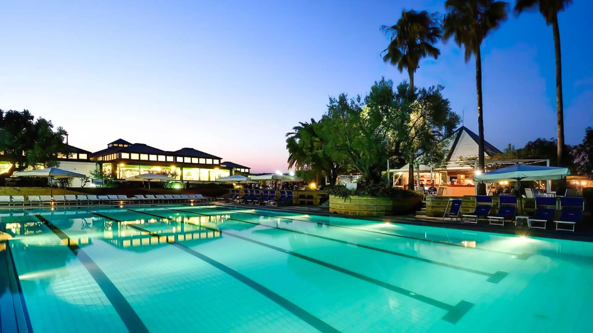 Piscina illuminata al crepuscolo con lettini, palme e edifici del resort sullo sfondo