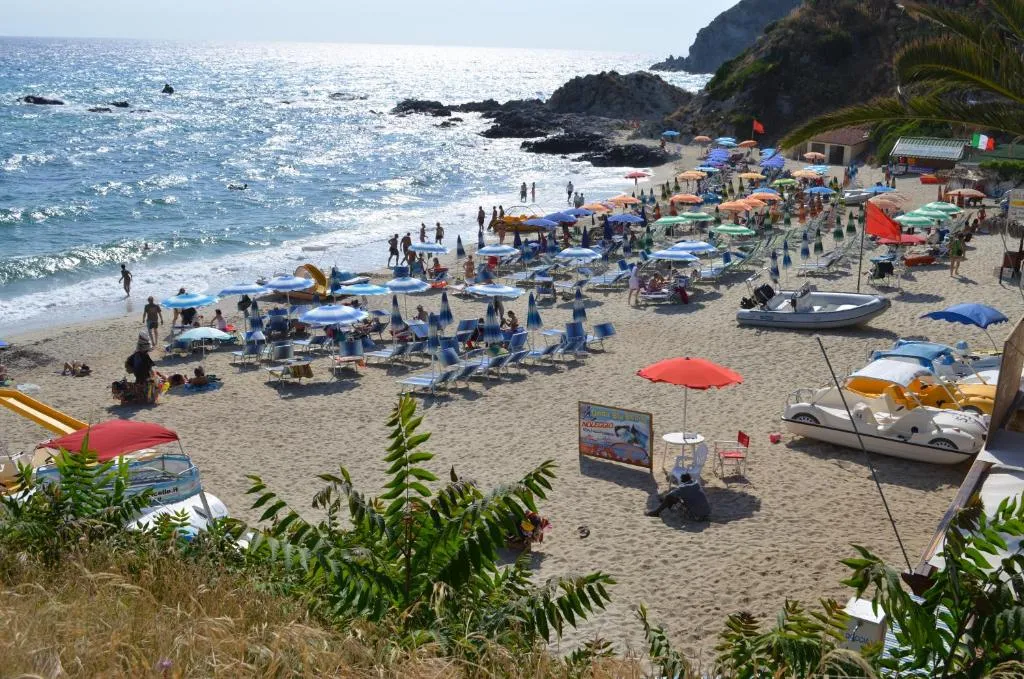 Spiaggia sabbiosa affollata con ombrelloni colorati, lettini e mare con scogli