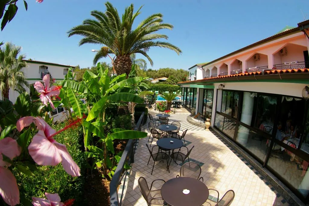 Giardino fiorito con tavoli, palme e piscina sullo sfondo dell'hotel