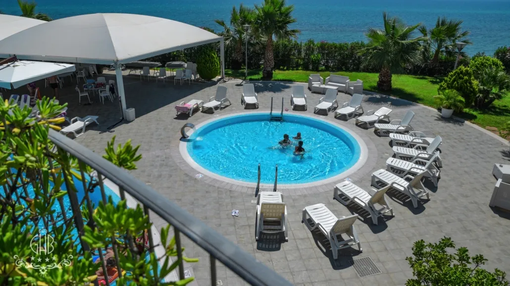 Piscina circolare con lettini bianchi e vista sul mare azzurro, circondata da palme