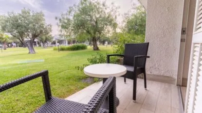 Balcone con due sedie nere in vimini e tavolino bianco, vista su giardino erboso con alberi.