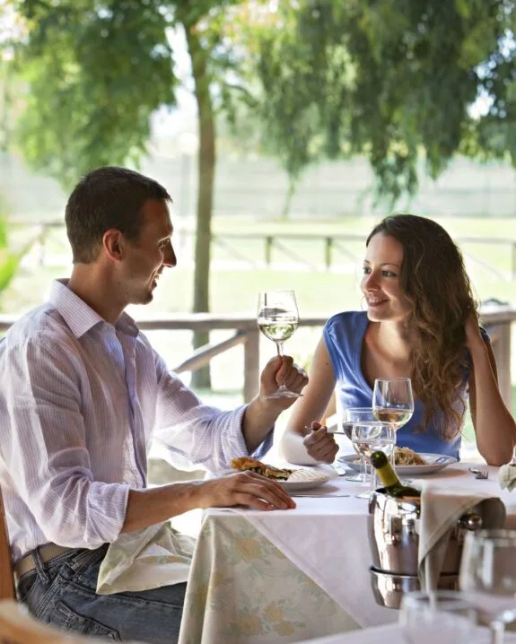 Coppia che brinda con calici di vino bianco durante un pranzo all'aperto in un ristorante con vista sul verde.