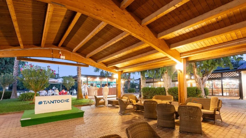 Area esterna coperta con sedute in vimini e tavoli, bar illuminato e persone in abiti bianchi sullo sfondo.
