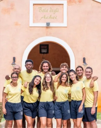 Gruppo di dieci giovani sorridenti con magliette gialle e pantaloncini blu davanti a un edificio beige con insegna "Baia degli Achei".