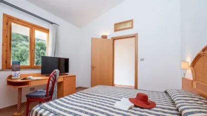 Camera da letto con letto matrimoniale a righe, scrivania con sedia rossa e finestra con vista sul verde esterno.
