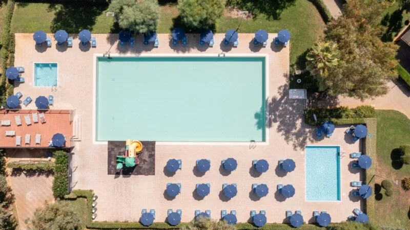 Vista aerea di piscina rettangolare con ombrelloni blu, lettini e area giochi per bambini in un giardino verde.