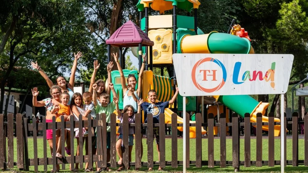 Bambini felici e due adulti davanti a un parco giochi colorato con scivoli e un cartello con scritta "TH Land!"