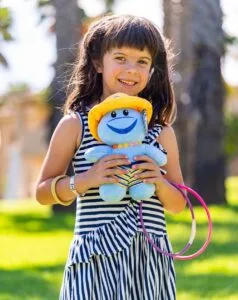 Bambina sorridente con vestito a righe che tiene un pupazzo azzurro con cappello giallo all'aperto su prato verde.