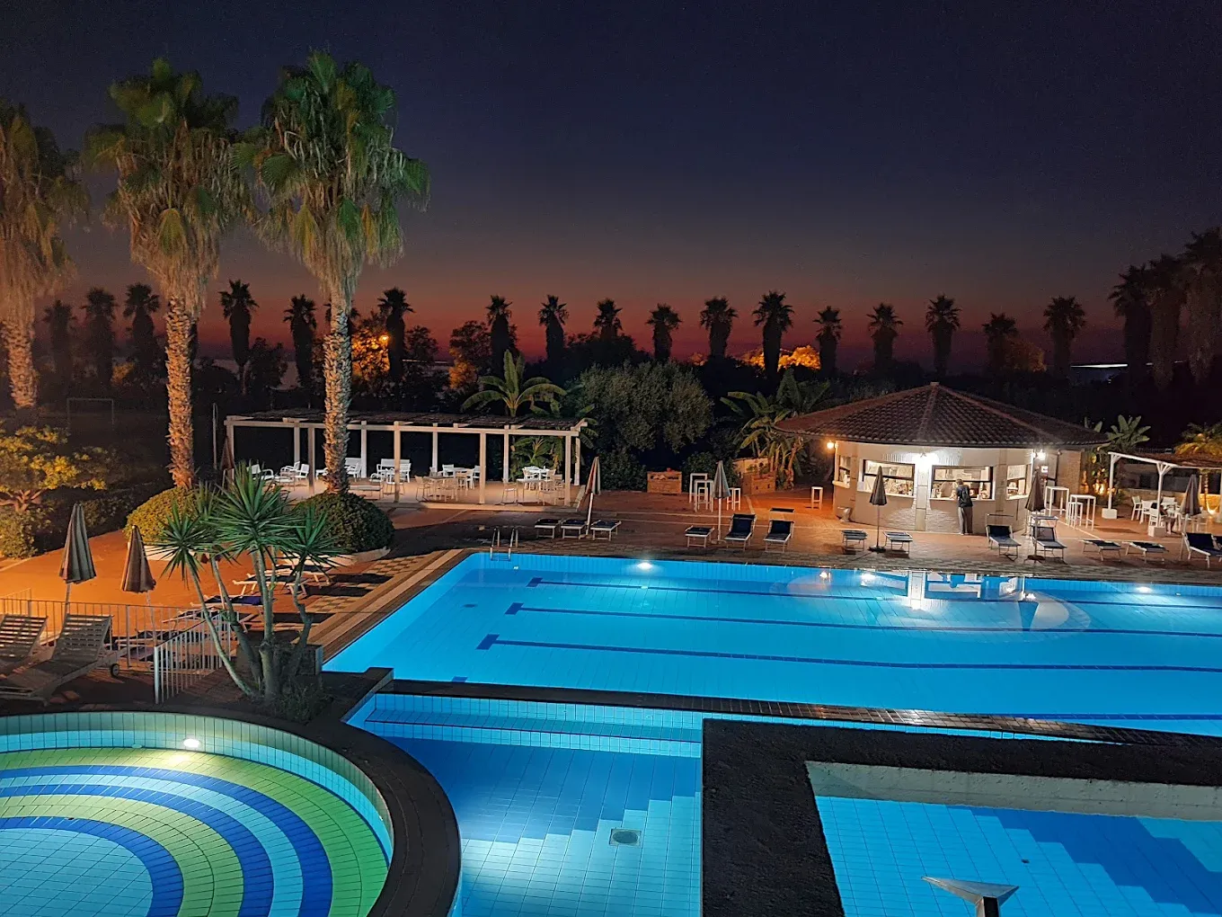 Piscine illuminate al tramonto con palme in controluce e bar all'aperto